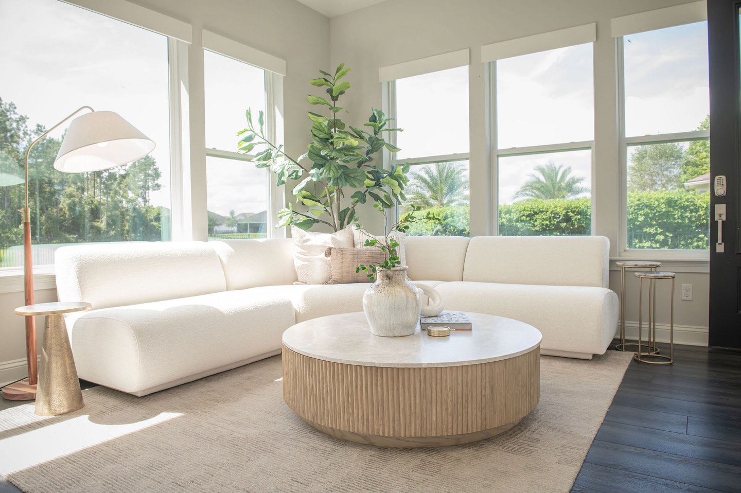 Sitting room with a curved cream boucle sectional facing a round white oak coffee table with a fluted base and stone top, a wall of windows looking onto palms and clipped hedges, a fiddle-leaf fig in a terracotta pot, a white arc floor lamp and slim brass nesting side tables
