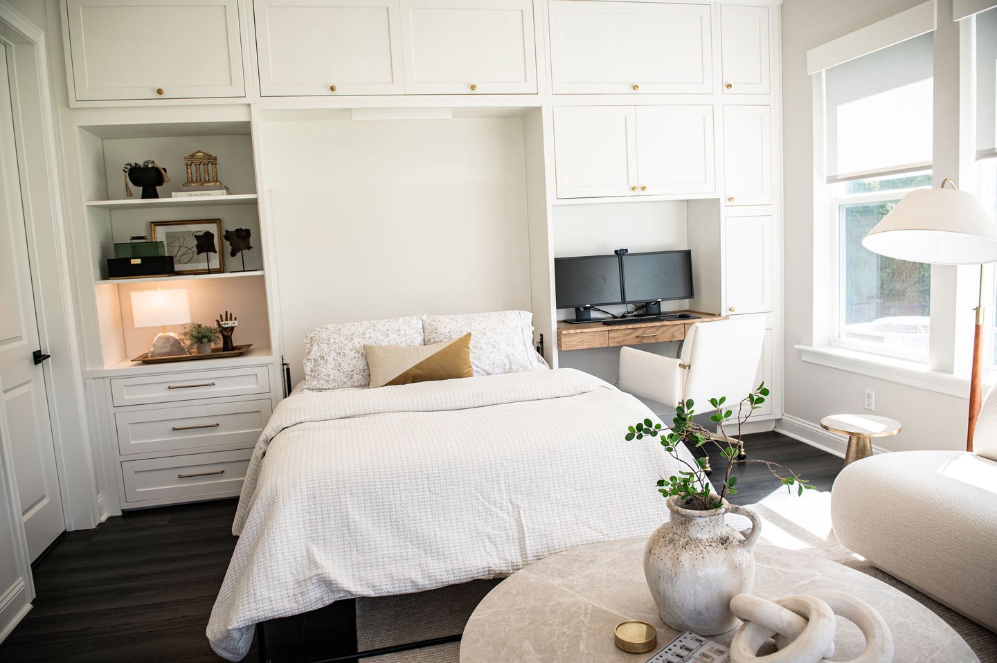 Guest room with a Murphy bed folded down and made up in white bedding with a mustard lumbar pillow, flanked by white built-in cabinetry, a lit display niche with a lamp and framed art, and a white oak desk with dual monitors and an upholstered chair beside a window