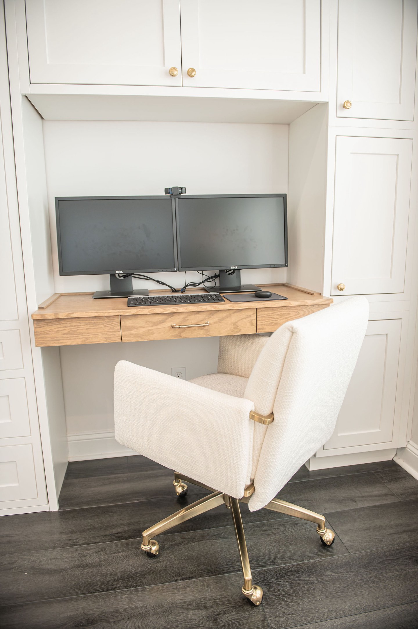 White oak fold-down desk set into a white built-in cabinet wall with brass hardware, holding two monitors and a keyboard, with a cream upholstered swivel chair on a brass base over dark wood flooring