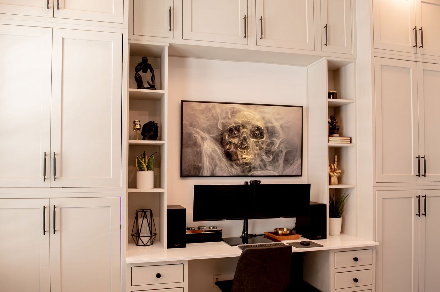 Floor-to-ceiling white shaker built-ins framing a recessed desk with a quartz top, dual monitors, a dark upholstered chair, open display niches holding sculptures, books and a potted plant, and a large framed smoke-and-skull canvas mounted above the desk