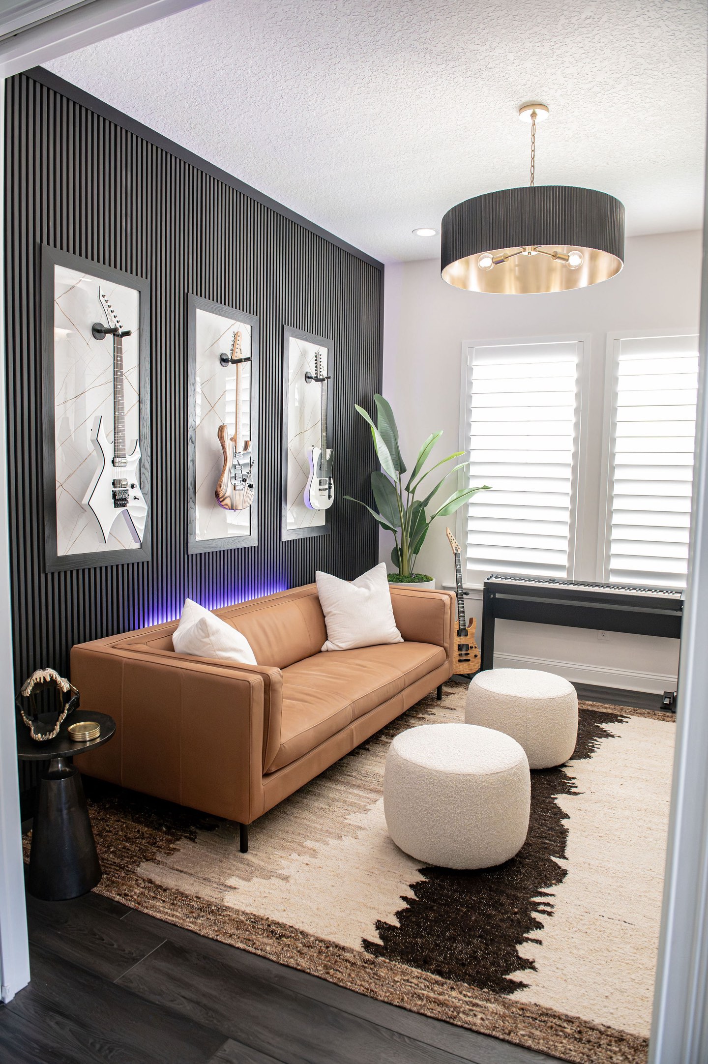 Home office with a black fluted wood accent wall hung with three framed electric guitars lit from below by violet LED strips, a cognac leather sofa with white cushions, two cream boucle poufs on a brown and cream hide-pattern rug, a black drum pendant with a brass interior, and plantation shutters beside a keyboard on a stand