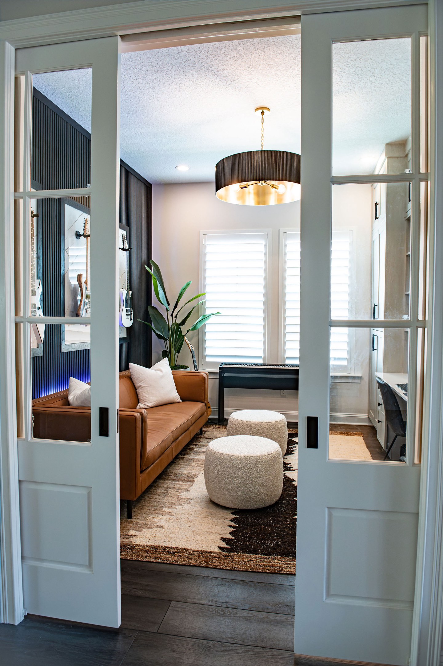 View through a pair of open white glass-paned pocket doors into a home office, showing the black fluted accent wall, a cognac leather sofa, two cream boucle poufs on a patterned hide rug and a black drum pendant with a brass interior