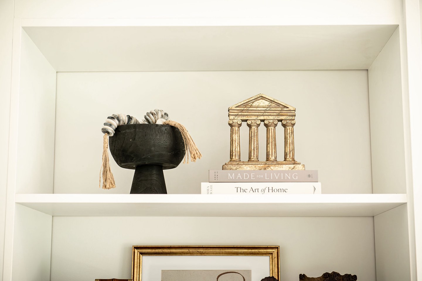 Close view of a white open shelf holding a black footed bowl with a tasseled throw, a small brass architectural model of a classical temple on a stack of design books, and a gilt-framed picture on the shelf below